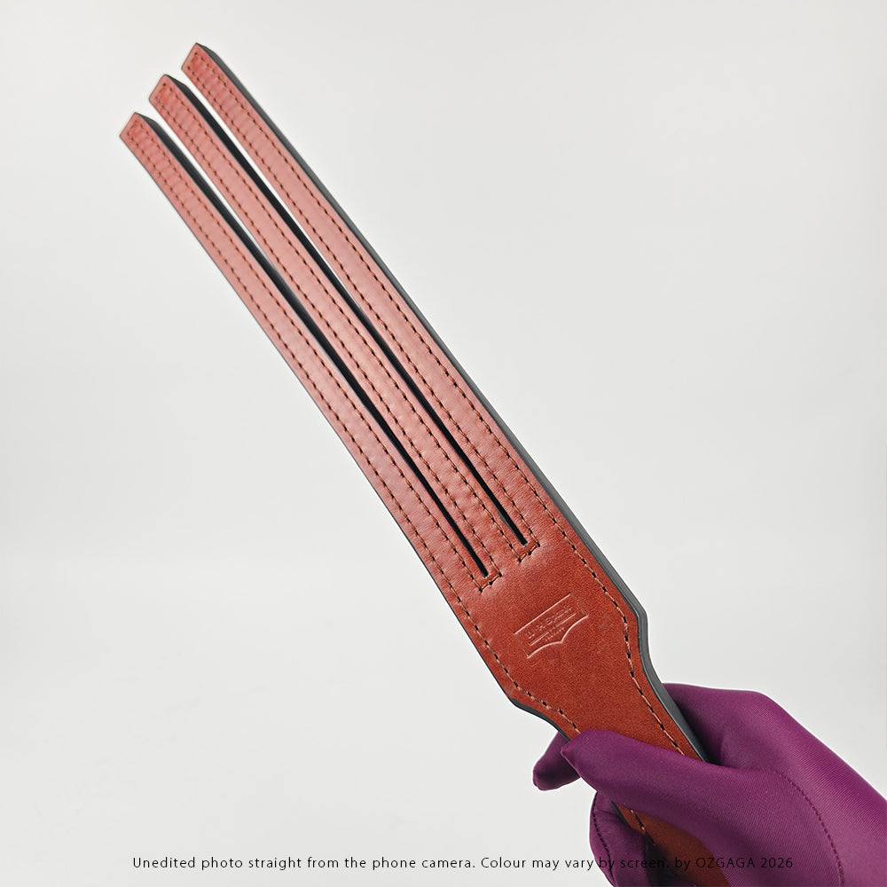 A close-up of a person's hand wearing a purple glove, holding a reddish-brown 3 Tailed Tawse against a light gray background.