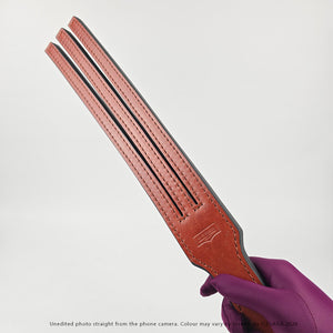 A close-up of a person's hand wearing a purple glove, holding a reddish-brown 3 Tailed Tawse against a light gray background.